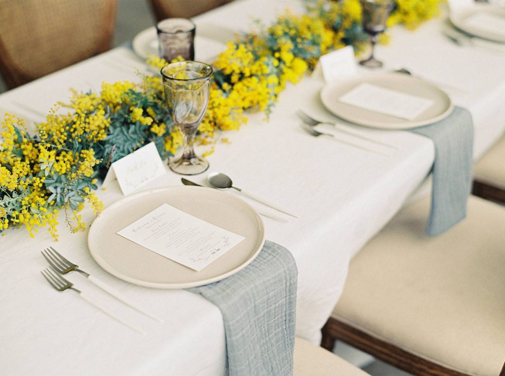 In questa foto un tavolo matrimonio con un runner di mimosa, tovagliato bianco e tovaglioli colore carta da zucchero posti sotto ad un piatto bianco