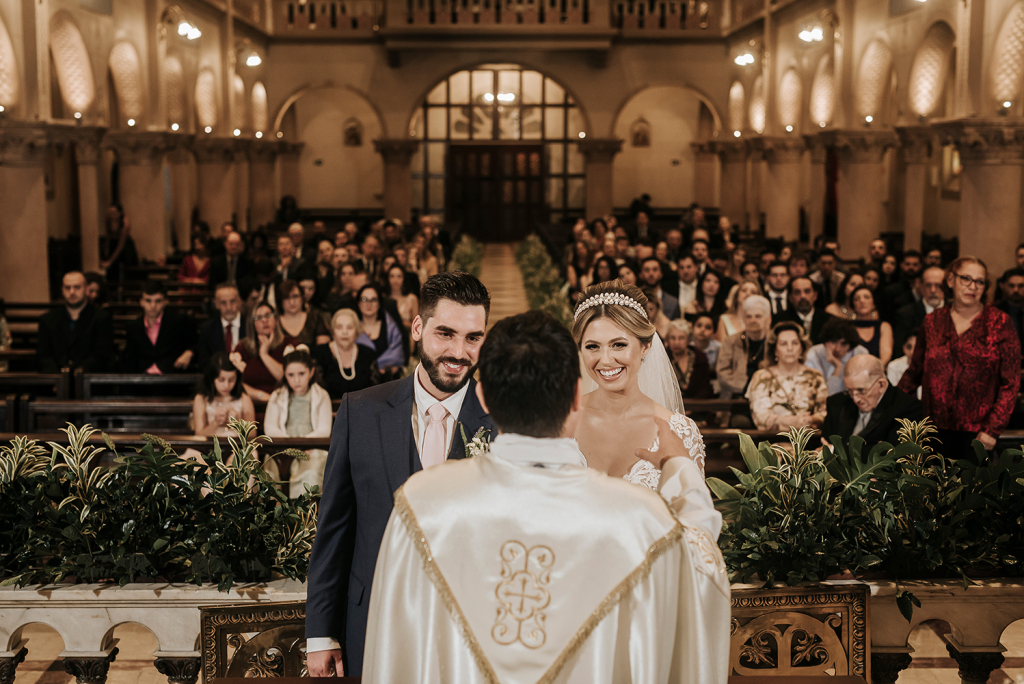 In questa foto due un matrimonio in chiesa. In primo piano è presente il sacerdote di spalle e davanti a lui sono ritratti gli sposi sorridenti. Dietro di loro, leggermente sfocati, tutti i loro ospiti. La sposa, bionda, porta tra i capelli un cerchietto di perle a cui è attaccato il velo. Lo sposo indossa una cravatta rosa