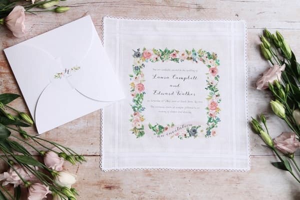 In quest'immagine, partecipazioni eleganti ricamate su un fazzoletto di stoffa con cornice floreale e busta di carta bianca con apertura a rosa