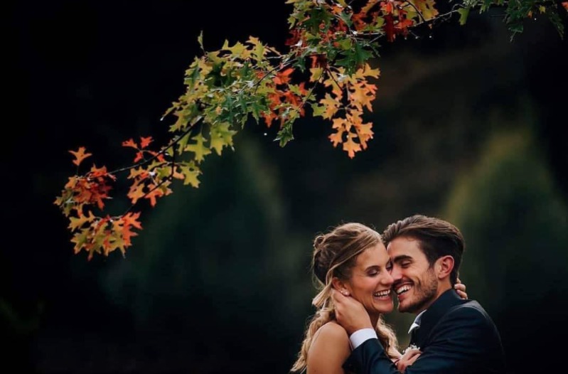 In questa foto uno sposa bacia sulla guancia la sua sposa sotto il ramo di un albero dai colori autunnali