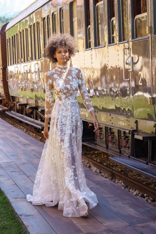 In questa foto una modella indossa un abito da sposa con le maniche 2022 della collezione Andrea Sedici. L'abito dal modello scivolato, con maniche lunghe, è totalmente arricchito da fiori ricamati.