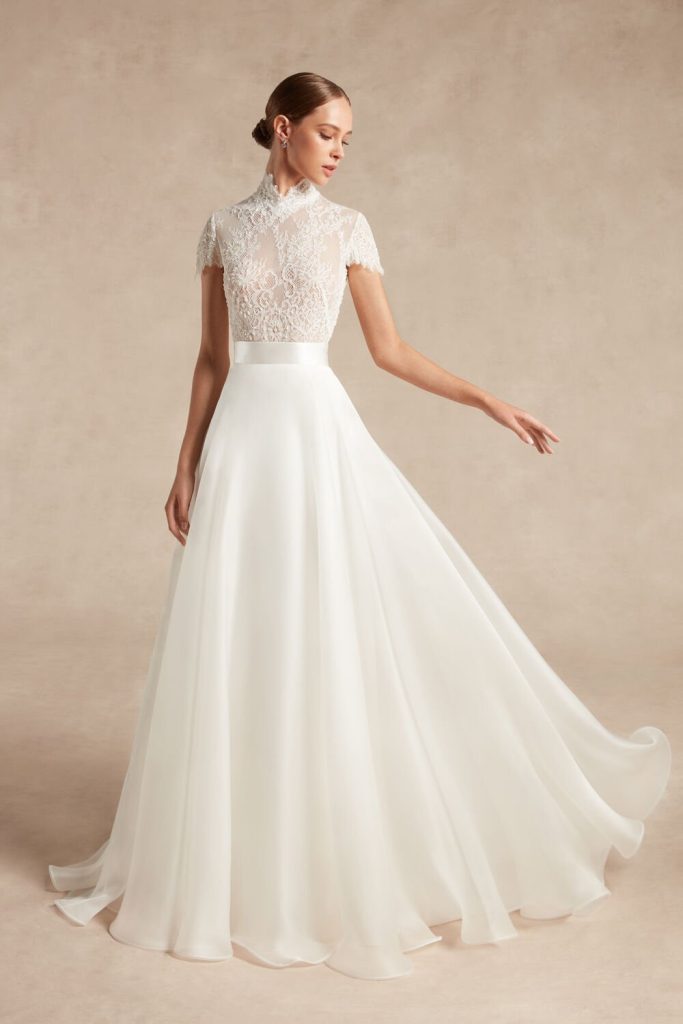 In questa foto una modella indossa un abito da sposa con le maniche 2022 della collezione Atelier Emè. L'abito a campana lungo, ha delle maniche corte di pizzo.