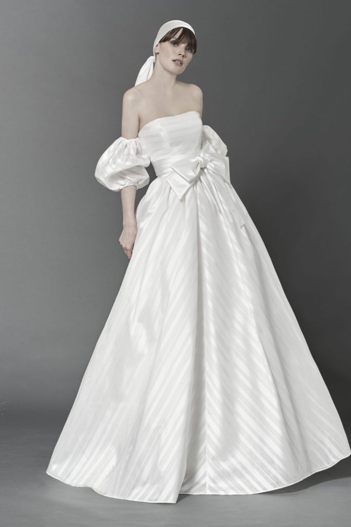 In questa foto una modella indossa un abito da sposa con le maniche 2022 della collezione Bellantuono. L'abito ampio con fiocco in vita, ha delle maniche rimovibili a palloncino.