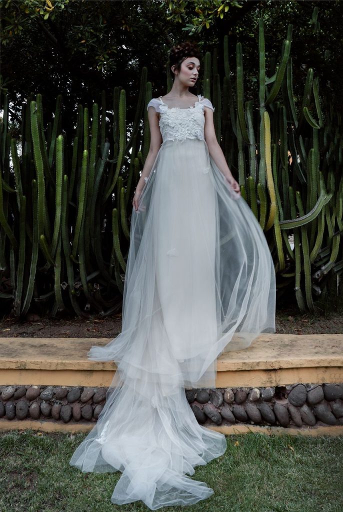 In questa foto una modella indossa un abito da sposa con le maniche 2022 della collezione Elisabetta Delogu. L'abito cade dritto con un corsetto riccamente decorato e le maniche corte di velo.