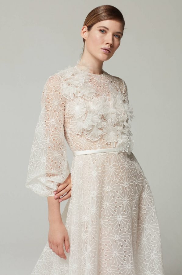 In questa foto una modella indossa un abito da sposa con le maniche 2022 della collezione Tosca Spose. L'abito è interamente ricoperto da un pattern di margherite trasparenti ed ha ampie maniche a sbuffo.