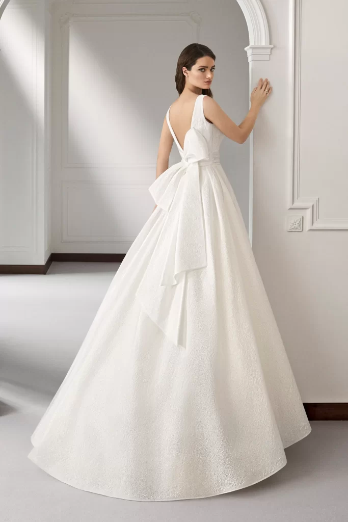 In questa foto una modella indossa un abito da sposa schiena scollata 2022 della collezione Atelier Emè con scollo profondo e maxi fiocco.