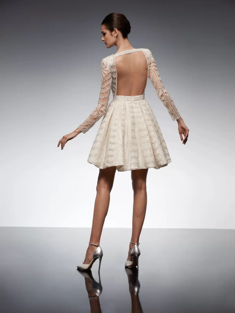 In questa foto una modella indossa un abito da sposa schiena scollata 2022 della collezione Nicole Milano con scollo profondo e maniche lunghe di pizzo.