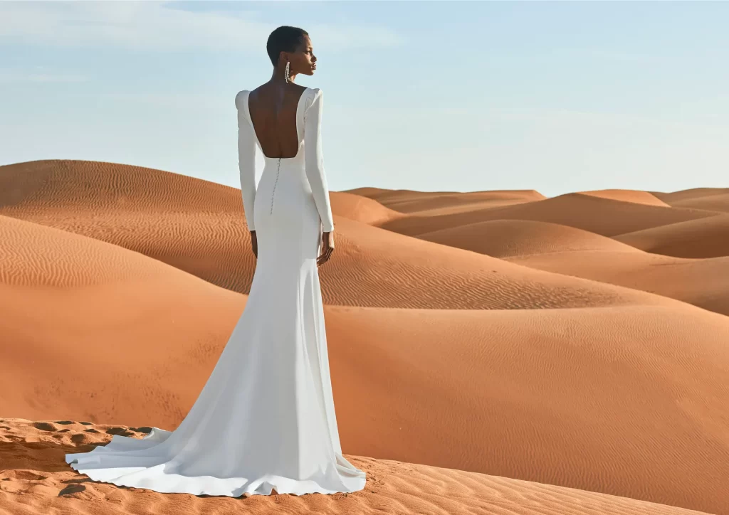 In questa foto una modella indossa un abito da sposa schiena scollata 2022 della collezione Pronovias con scollo profondo e maniche.