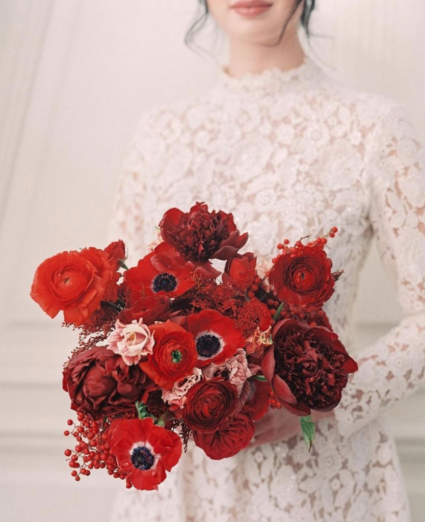 In questa foto una sposa ripresa dal collo in giù con un bouquet di anemoni e peonie rossi. La sposa indossa un abito in pizzo macramè
