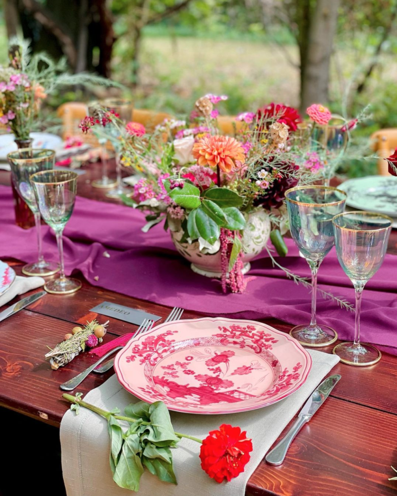 In questa foto il dettaglio di un tavolo di legno nudo decorato con un runner fucsia su cui sono poggiati vasetti di fiori di campo rosa, rossi e arancioni. In primo piano è inquadrato un tovagliolo colore salvia su cui è poggiato un piatto rosa e fucsia con davanti un garofano rosso 