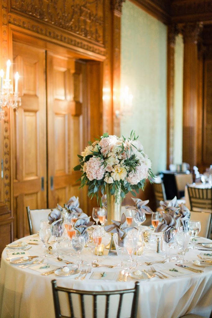 In questa foto un tavolo matrimonio rotondo con un centrotavola composto da un vaso colore oro con fiori rosa e colore giallo tenue. Sul tavolo i tovaglioli sono inseriti nei calici