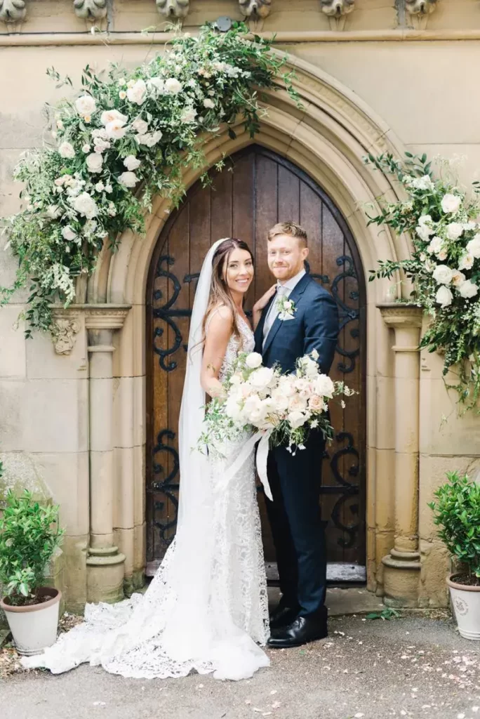 In questa foto due sposi posano sorridenti davanti al portone chiuso della chiesa in cui è stato celebrato il loro matrimonio, decorato con fiori matrimonio bianchi e foglie. La sposa tiene nella mano destra un bouquet a braccio di peonie bianche