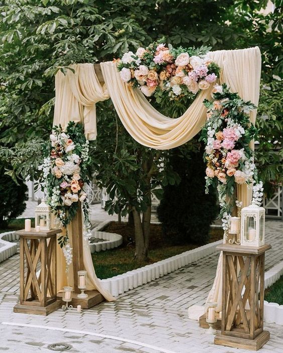 In questa foto una arco di legno usato come ingresso ad un matrimonio civile decorato con drappi di lino colore panna e composizioni di foglie e fiori per matrimonio rosa, bianchi e colore pesca. Alla base sono presenti due alzate con candele e lanterne bianche 