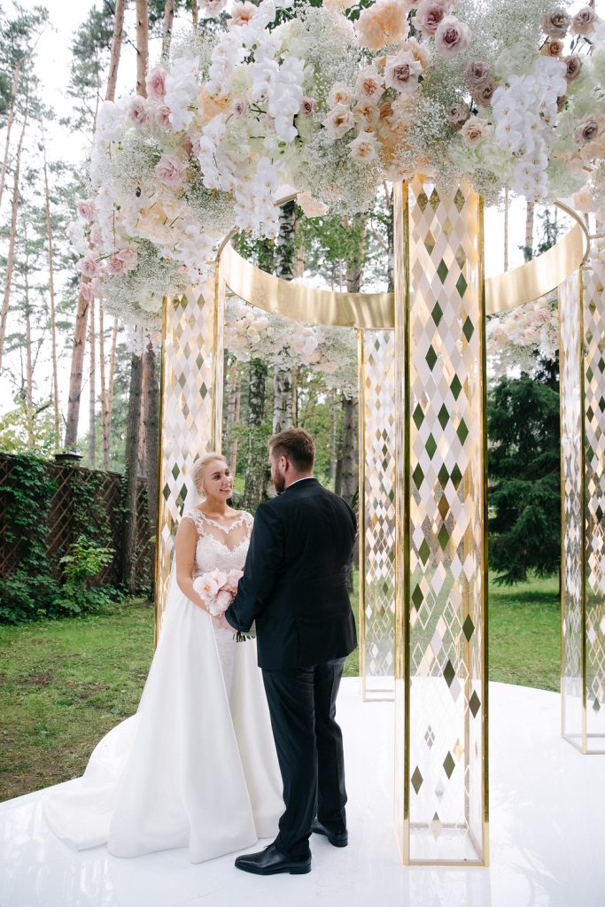 In questa foto due sposi mano nella mano sotto ad un gazebo colore oro circolare decorato in cima con fiori matrimonio bianchi, rosa e nebbiolina