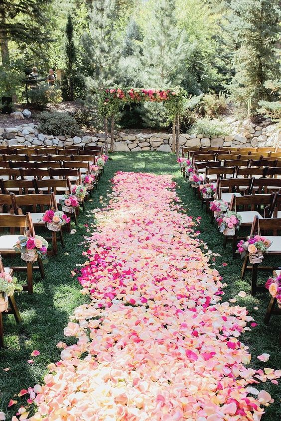 In questa foto la location di un matrimonio civile su un prato. Il corridoio centrale è ricoperto di petali rosa, bianchi e fucsia. Sulle sedie sono legati mazzolini di fiori abbinati. Alla fine del corridoio centrale è presente un arco di legno con fiori rosa, bianchi e fucsia