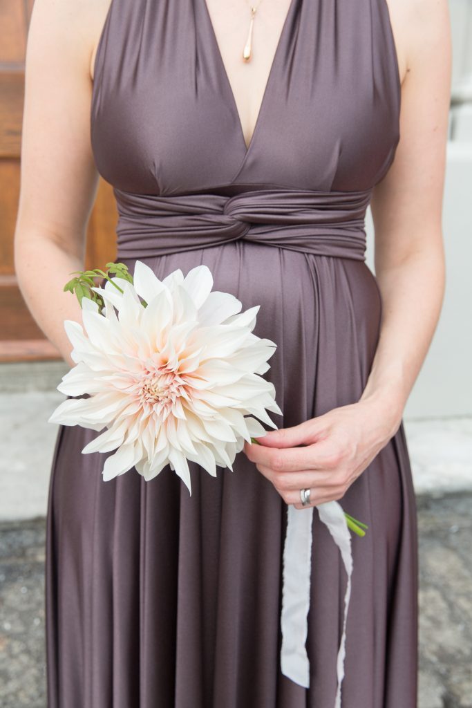 In questa foto una damigella inquadrata dal busto in giù tiene tra le mani un fiori di dalia rosa. Indossa un abito colore melanzana con scollo all'americana