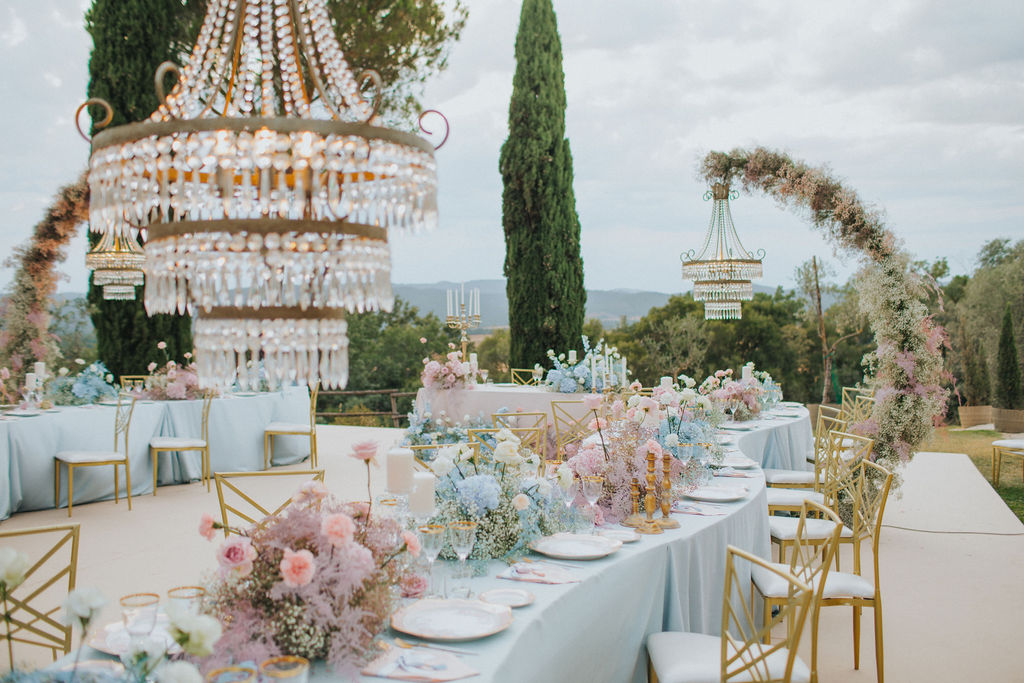 In questa foto l'allestimento di un ricevimento con centrotavola alternati di fiori per matrimonio nei colori celeste e rosa. I tavoli usati sono lunghi disposti a forma di S con tovagliato azzurro. Accanto ai tavoli sono posizionati: sedie colore oro e bianche e delle strutture ricoperte di fiori bianchi e rosa a cui sono appesi lampadari in cristallo 