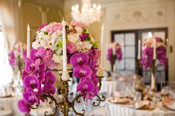 In questa foto in primo piano uno dei centrotavola della sala di un ricevimento di nozze composta da un candelabro colore bronzo, orchidee viola pendenti, peonie rose e orchidee bianche