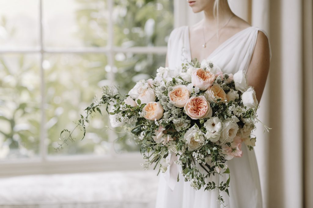 In questa foto una sposa inquadrata dalla bocc in giù tiene tra le braccia un bouquet di rose inglesi colore cipria e bianco e foglie 