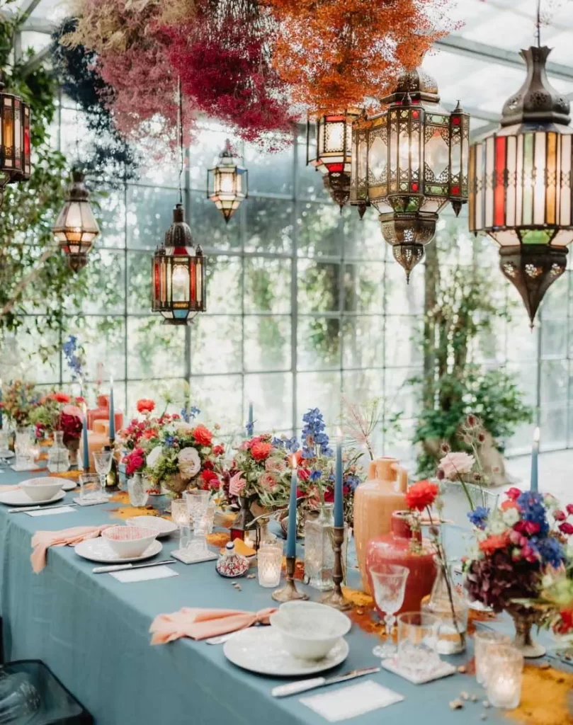 In questa foto un tavolo matrimonio allestito in una serra. Sopra al tavolo sono disposti fiori secchi nei colori dell'arancione, del bordeaux, rosa, bianco e blu, e lampadari colorati in stile orientale. Il tavolo è decorato con tovaglia colore avion, tovaglioli rosa e tanati centrotavola con fiori di campo per matrimonio colorati, candele e vasi 