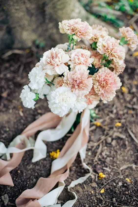 In questa foto un bouquet di garofani rosa cipria e bianchi fermati da due nastri di colore rosa e bianco, poggiato sul terreno in un bosco