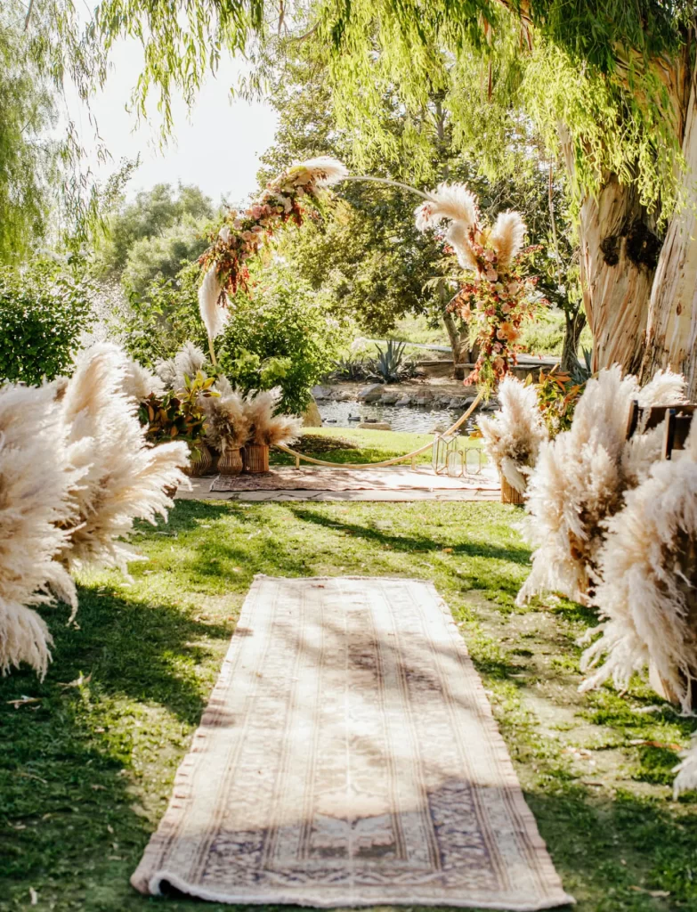 In questa foto la location per un matrimonio civile allestito in stile boho chic con pampas e un tappeto lungo il corridoio. L'altare è allestito con un cerchio decorato con fiori e pampas