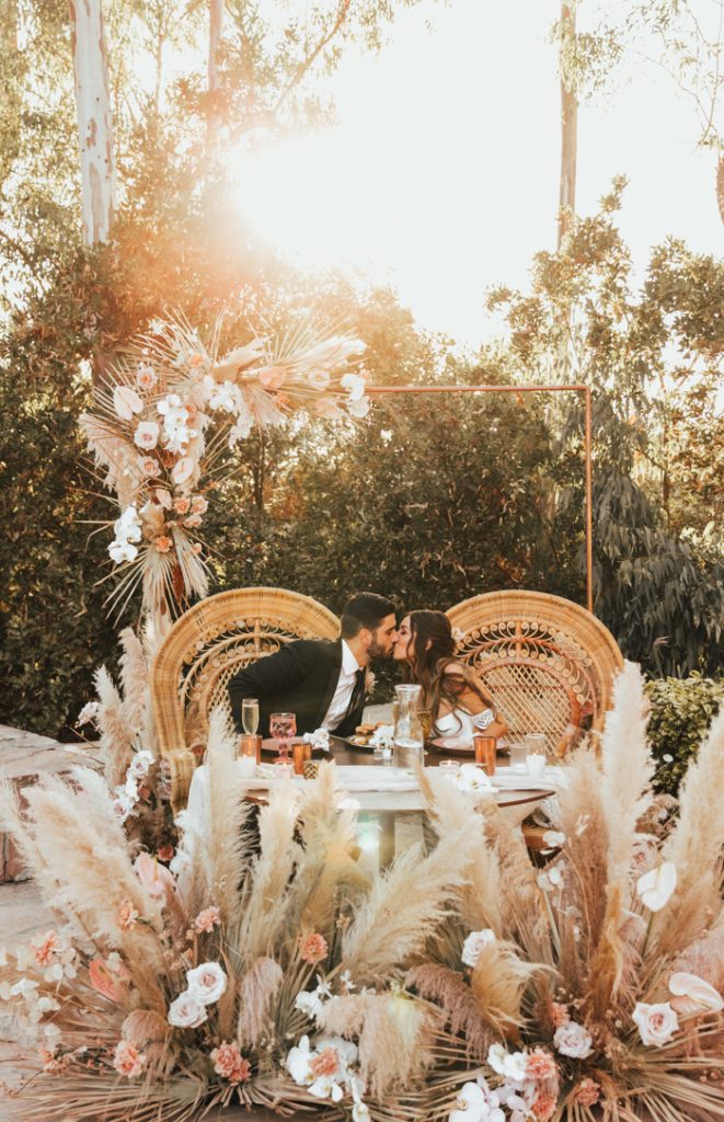 In questa foto due sposi seduti al loro tavolo su sedie in vimini circondati da pampas, rose bianche e rosa. Dietro di loro è presente una cornice oro rettangolare decorata con palme e fiori bianchi