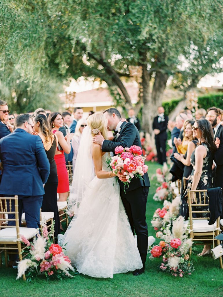 In questa foto due sposi si baciano dopo la celebrazione del loro matrimonio civile circondati dai loro invitati e dai loro fiori matrimonio rosa e bianchi. La sposa tiene in mano un bouquet di peonie fucsia e rosa