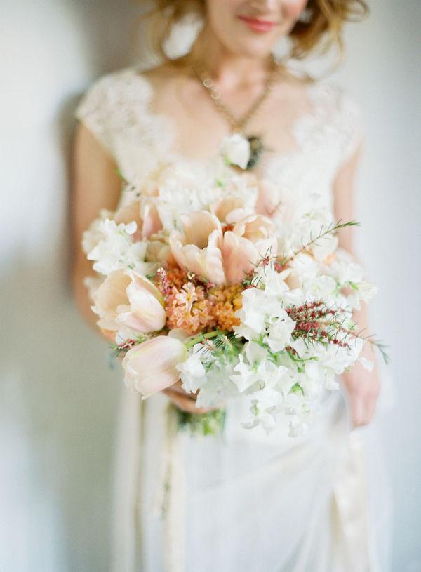 In questa foto una sposa bionda inquadrata dalle labbra in giù mentre tiene nella mano destra un bouquet di tulipani rosa cipria e fiori matrimonio bianchi