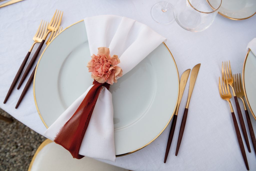 In questa foto il dettaglio di un segnaposto con un garofano rosa legato ad un nastro rosso pendente sul tovagliolo bianco legato al piatto. Accanto al patto con bordatura colore oro sono disposte posate bordeaux e colore oro