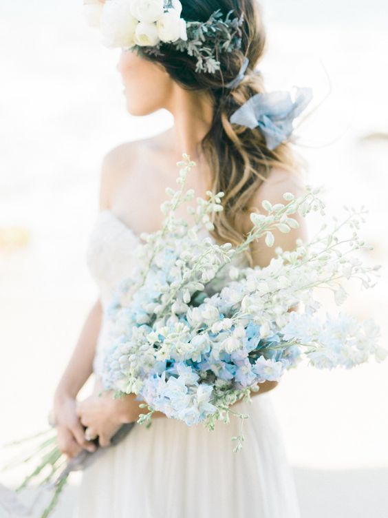 In questa foto una sposa di spalle che tiene tra le braccia un bouquet di delphinium azzurri e bianchi. Tra i capelli porta una coroncina di fiori matrimonio bianchi e azzurri