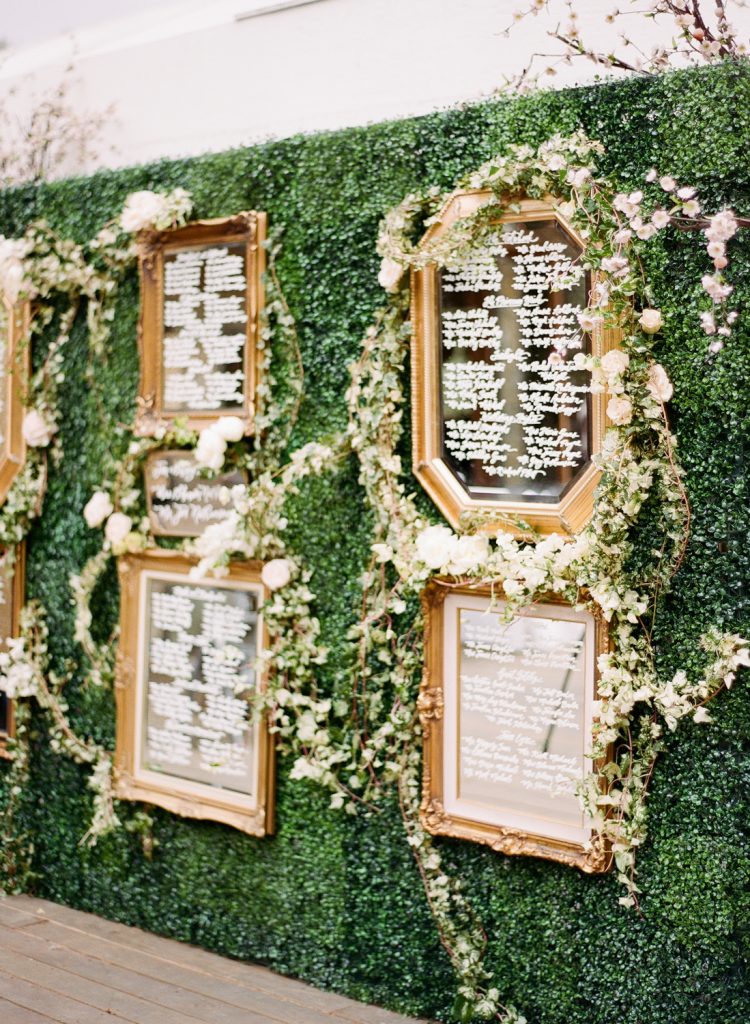In questa foto un tableau de mariage su un muro di foglie e fiori bianchi con i nomi degli invitati scritti con un pennarello bianco su specchi circondati da cornici colore oro