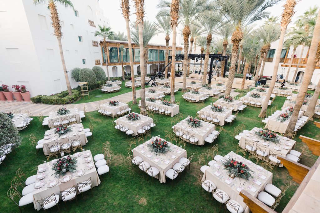 In questa foto i tavoli quadrati di un ricevimento di nozze su un prato tra le palme decorati con fiori fucsia e foglie su tovagliato beige