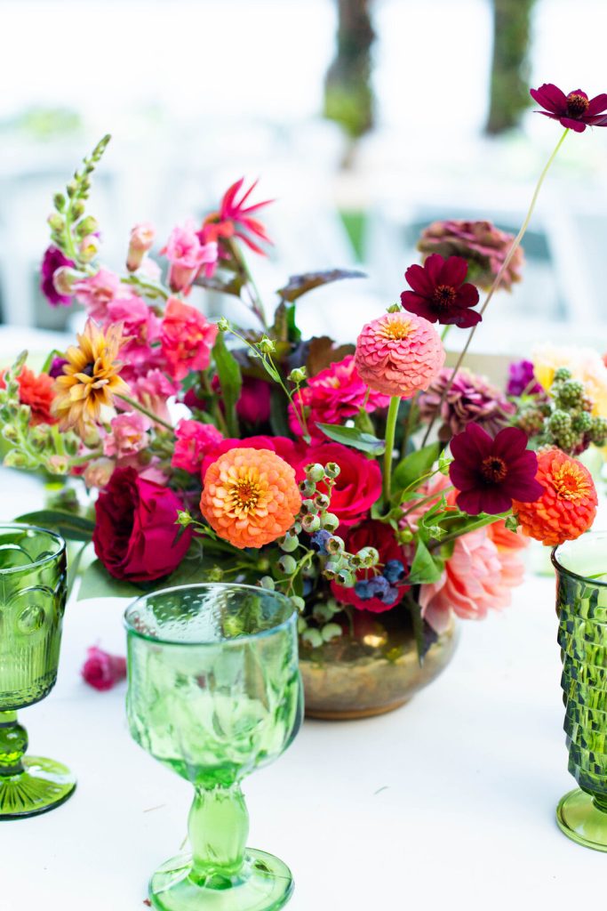 In questa foto un centrotavola con fiori di zinnia arancione e rosa, rose rosse e fiori di campo colorati contenuti in un vaso bronzo tra bicchieri verdi 