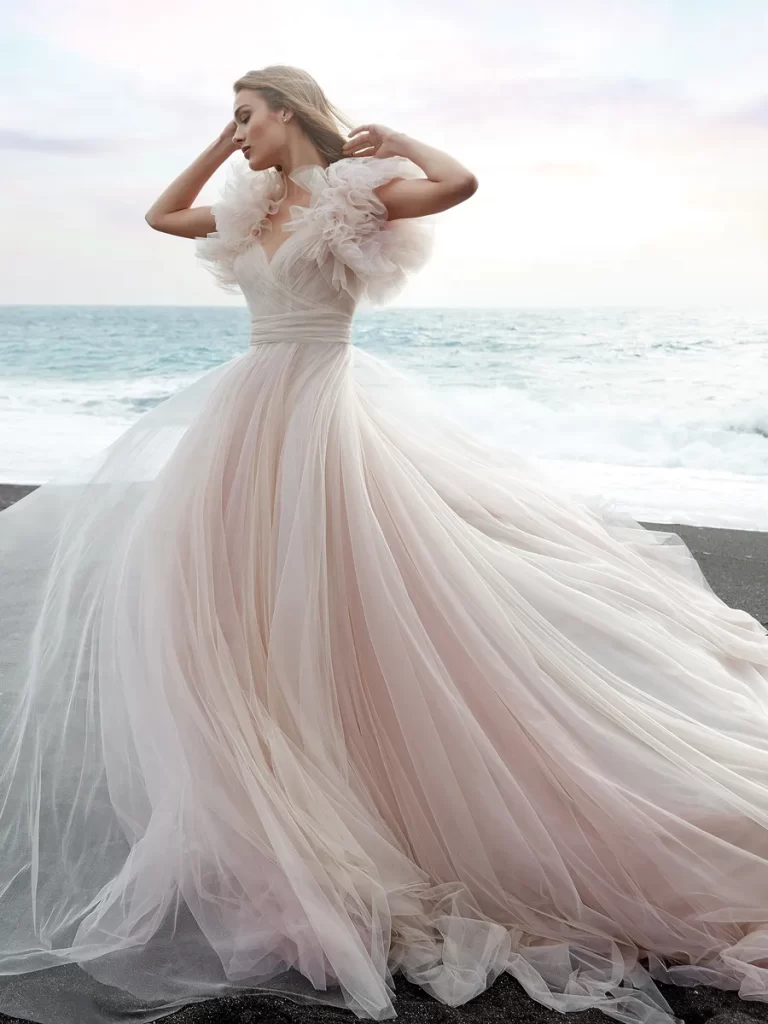 In questa foto una modella indossa un abito da sposa principessa Nicole Milano posando su una spiaggia con lo sfondo del mare