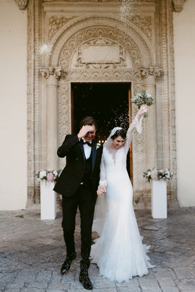 In questa foto due sposi felici si tengono per mano mentre escono dalla chiesa durante il lancio del riso. La sposa tiene in alto il bouquet con la mano sinistra, mentre lo sposo si copre la fronte con la mano destra