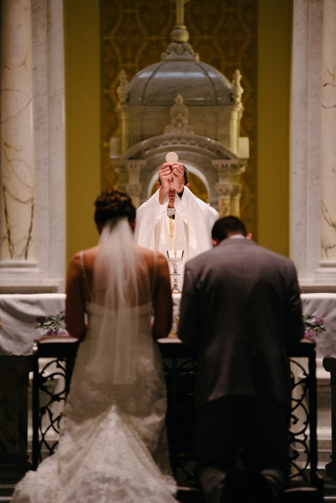 In questa foto due sposi che pregano di spalle in chiesa mentre il sacerdote solleva l'Eucarestia