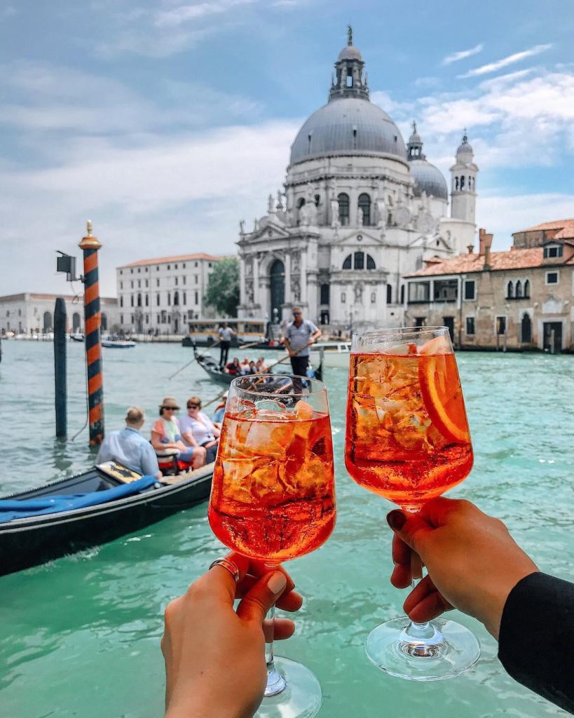 In questa foto le mani di due sposi che brindano con un Aperol Spritz a Venezia durante il loro viaggio di nozze. Sullo sfondo si vede gondole e gondolieri che navigano in un canale
