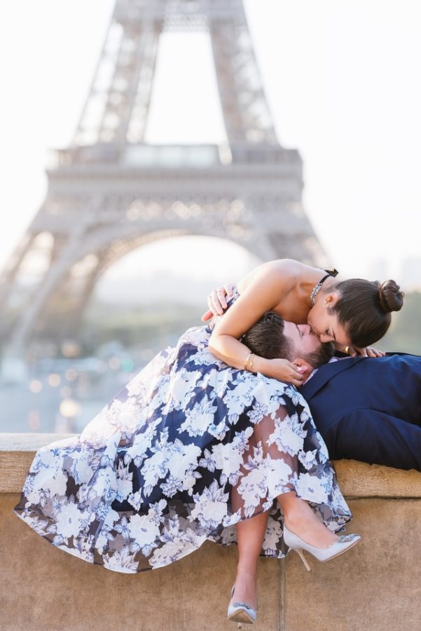 Una coppia si bacia sdraiati su un muretto davanti alla Torre Eiffel durante il loro viaggio di nozze a Parigi. Lei indossa un abito nero e bianco e fiori e una paio di décolleté Manolo Blahnik colore grigio perla e porta i capelli acconciati in uno chignon