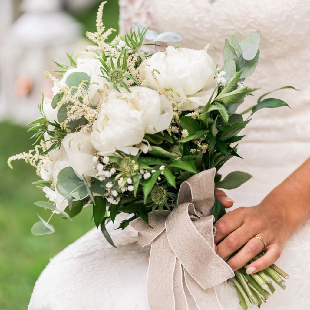 In questa foto le mani di una sposa che tiene poggiato sulle gambe un piccolo bouquet scomposto di peonie bianche, eucalipto e foglie di felce stretti con un nastro di lino beige