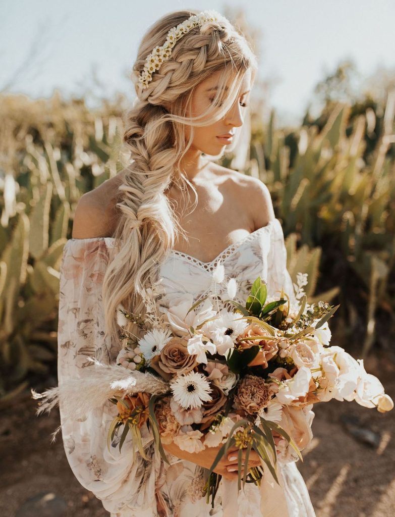 In questa foto un'acconciatura da sposa boho chic con una grande treccia laterale decorata con una fascia di piccole margherite. La sposa  tiene tra le mani un bouquet scomposto nei toni del bianco e e del beige