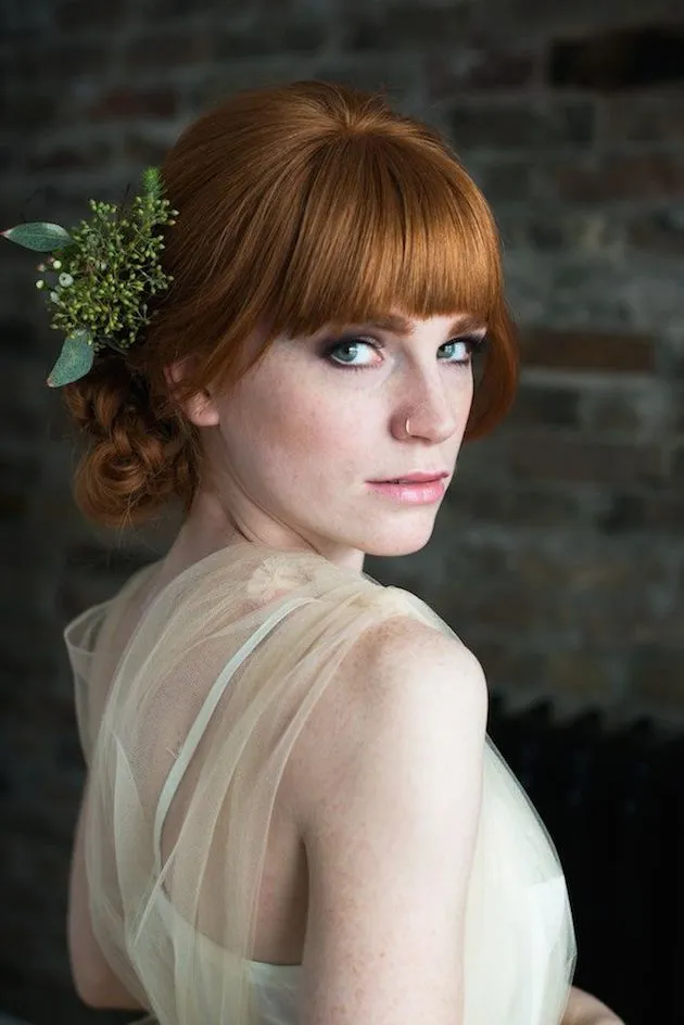 In questa foto una sposa con capelli rossi porta un'acconciatura semiraccolta con frangia e decorata con nebbiolina