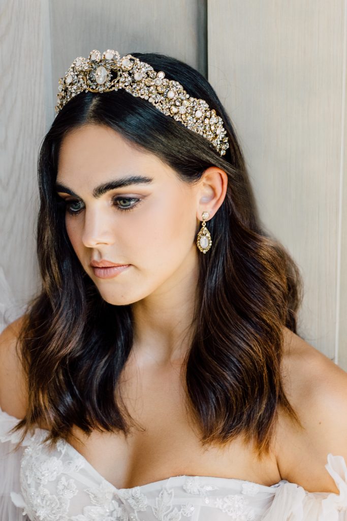In questa foto una modella con capelli medi sciolti indossa una corona da sposa colore oro con cristalli e orecchini pendenti abbinati 