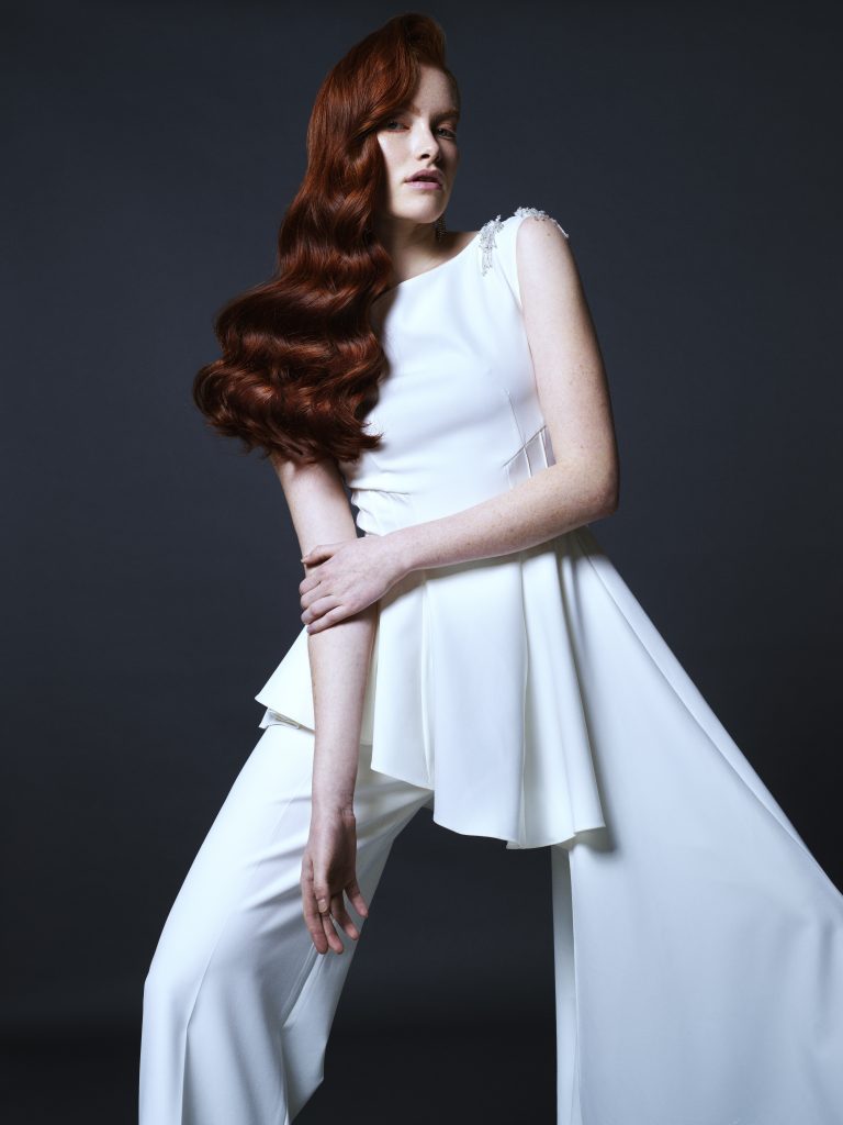 In questa foto una modella con i capelli rossi sciolti e mossi posa con un abito da sposa con pantaloni e blusa