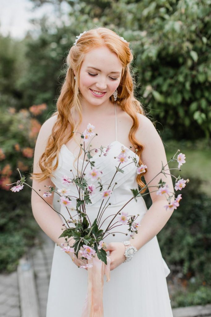 In questa foto una sposa con viso tondo porta un'acconciatura semiraccolta e, guardando verso il basso, tiene tra le mani un bouquet di fiori di ciliegio