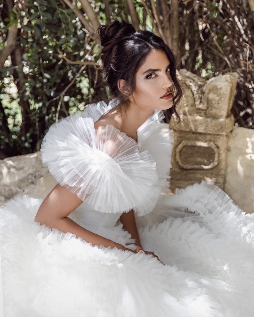 In questa foto una modella con un'acconciatura da sposa con uno chignon morbido e ciocche di capelli scomposte sul viso. Indossa un abito con ampie rouches in tulle sulle spalle e posa tra gli alberi davanti ad una fontana in pietra 