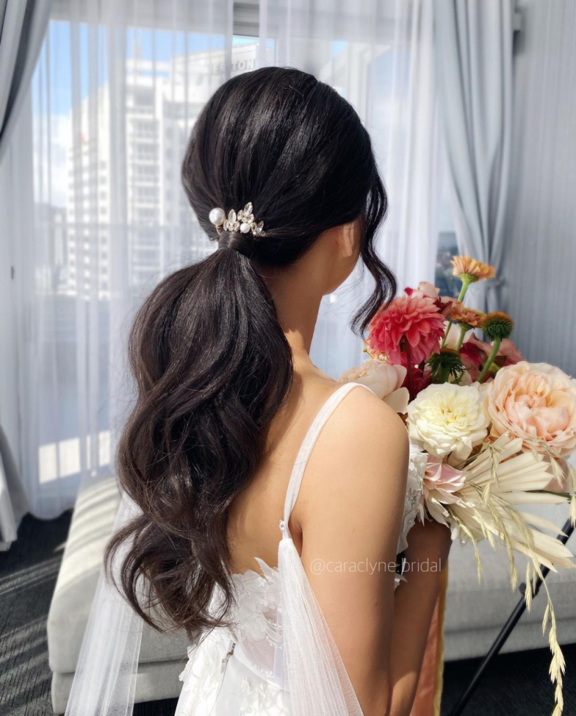 In questa foto una sposa con una grande coda di capelli bassa e decorata con un piccolo fermaglio di colore bianco all'attaccatura. Tiene nelle mani un bouquet di peonie di colore rosa e bianco