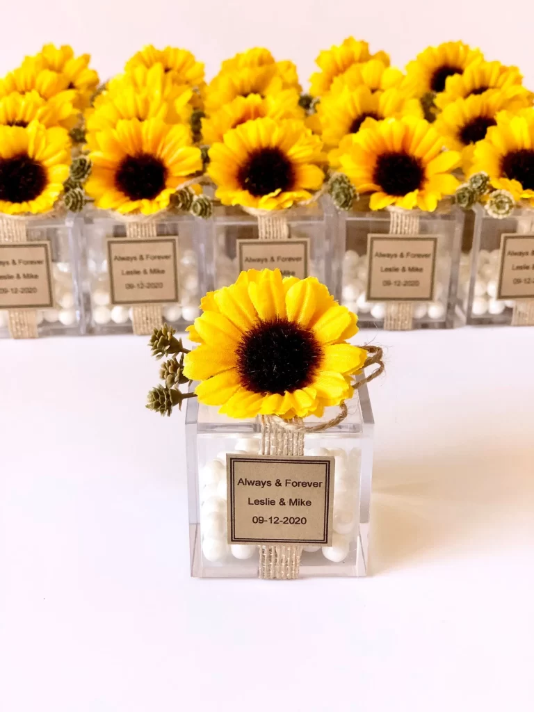 In questa foto bomboniere per matrimonio con girasoli a forma di scatoline in plexiglass con fiore sulla chiusura e bigliettino con nome degli sposi e data delle nozze