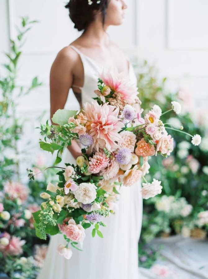 In questa foto una sposa con abito scivolato tiene nella mano destra un bouquet a casacata con rose bianche, dalie e fiori di zinnia rosa, fiori lilla, margherite e foglioline