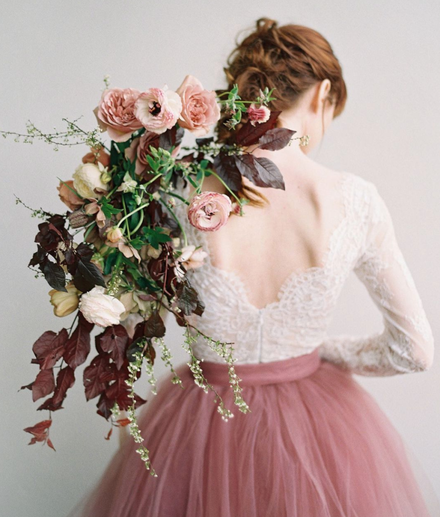 In questa foto una modella di spalle con gonna in tulle rosa e body di pizzo bianco tiene sulla spalla sinistra un bouquet a cascata nei toni del rosa e borgogna con rose inglesi e foglie d'edera viola